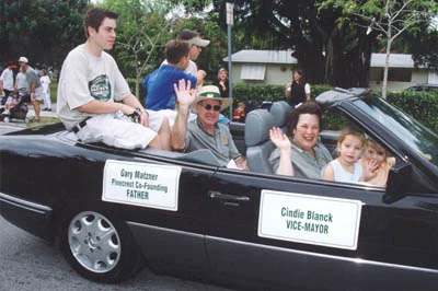 Gary Matzner waving from a convertible in a Pinecrest parade, with a sign reading “Gary Matzner, Pinecrest Co-Founding Father.”